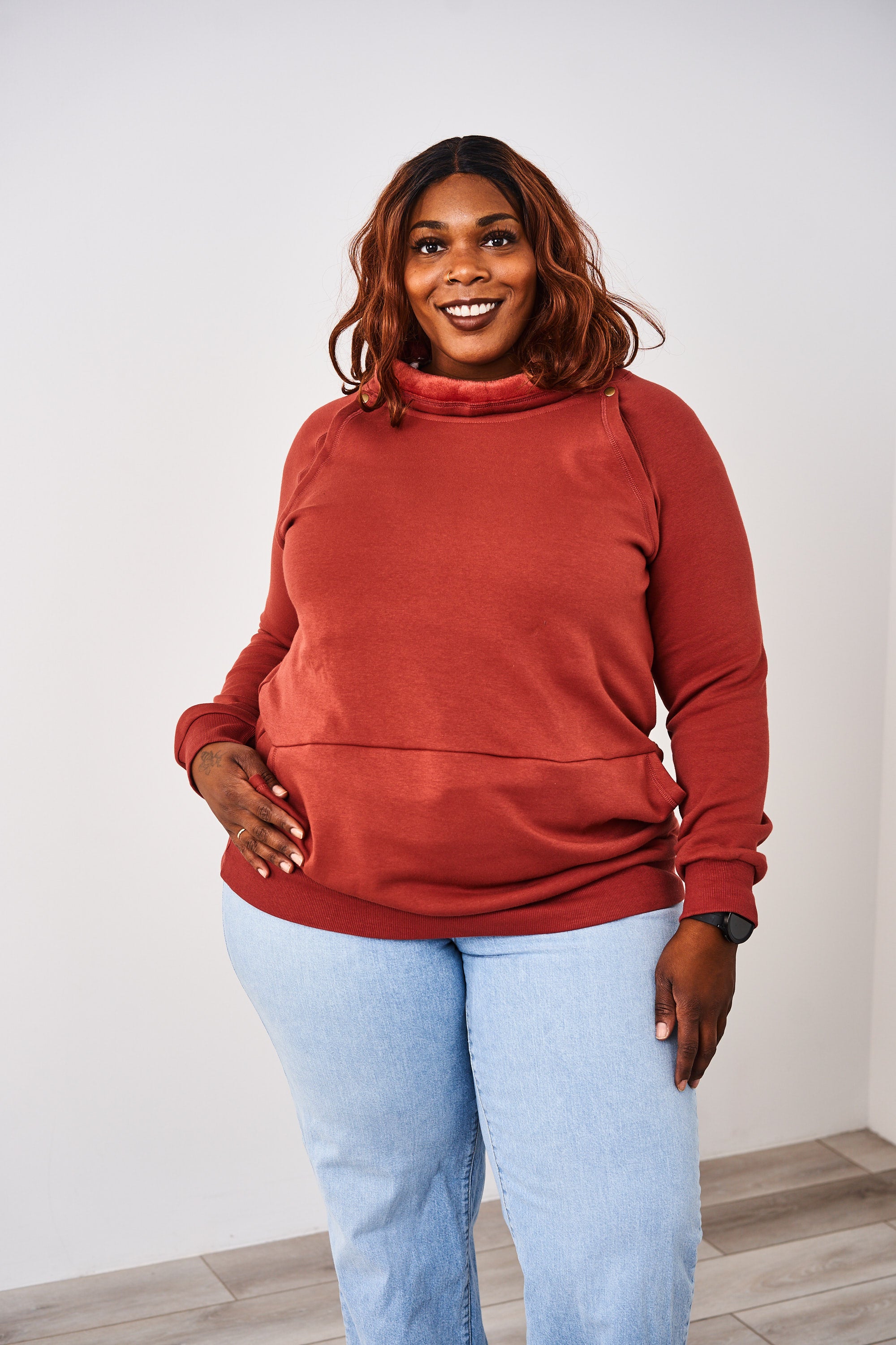 Woman wearing a Latched Mama Rust Harbor Snap Nursing Pullover with a white background and wood floor