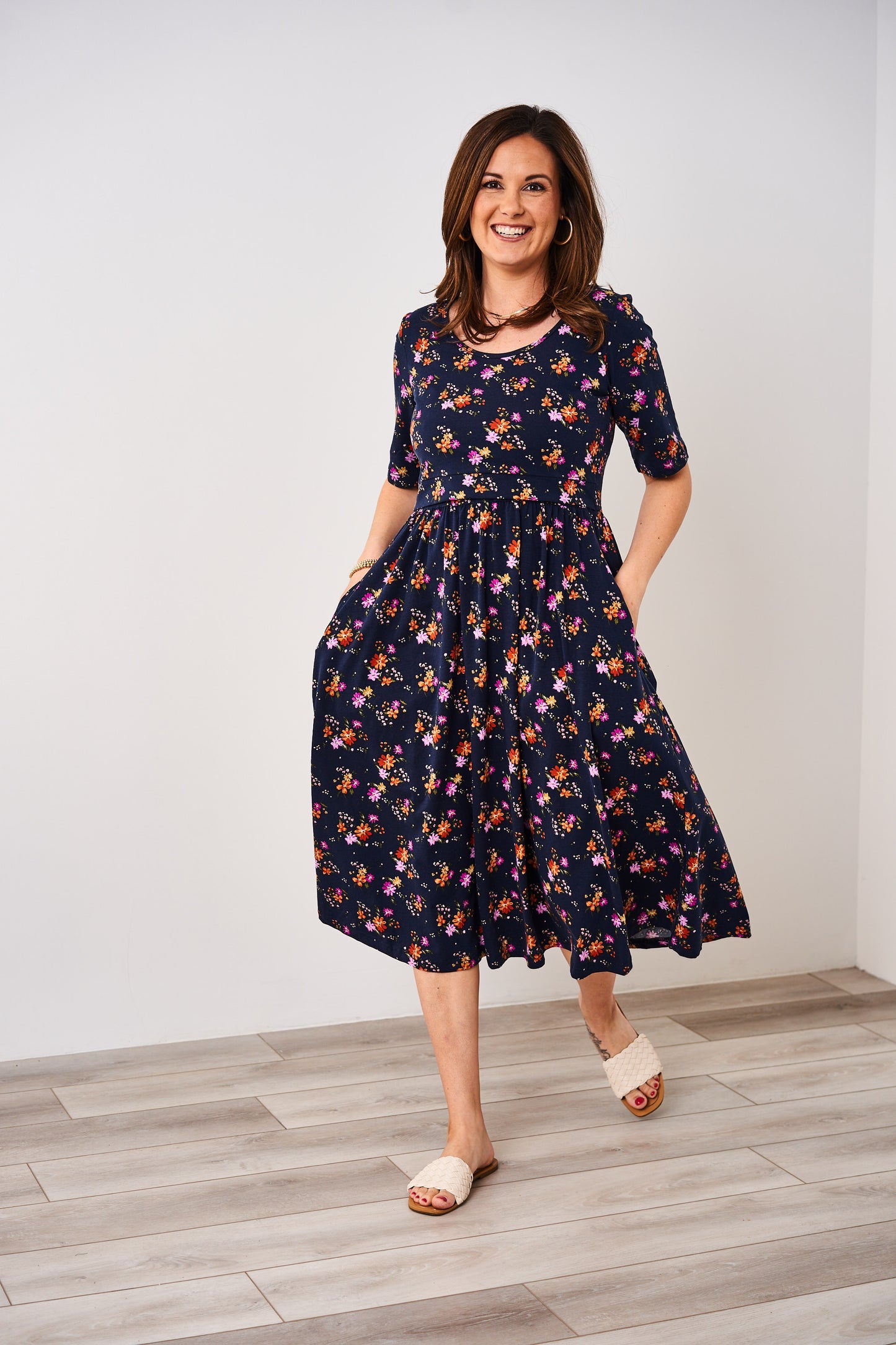 Woman wearing a Latched Mama floral nursing dress standing on a wooden floor with a white wall background