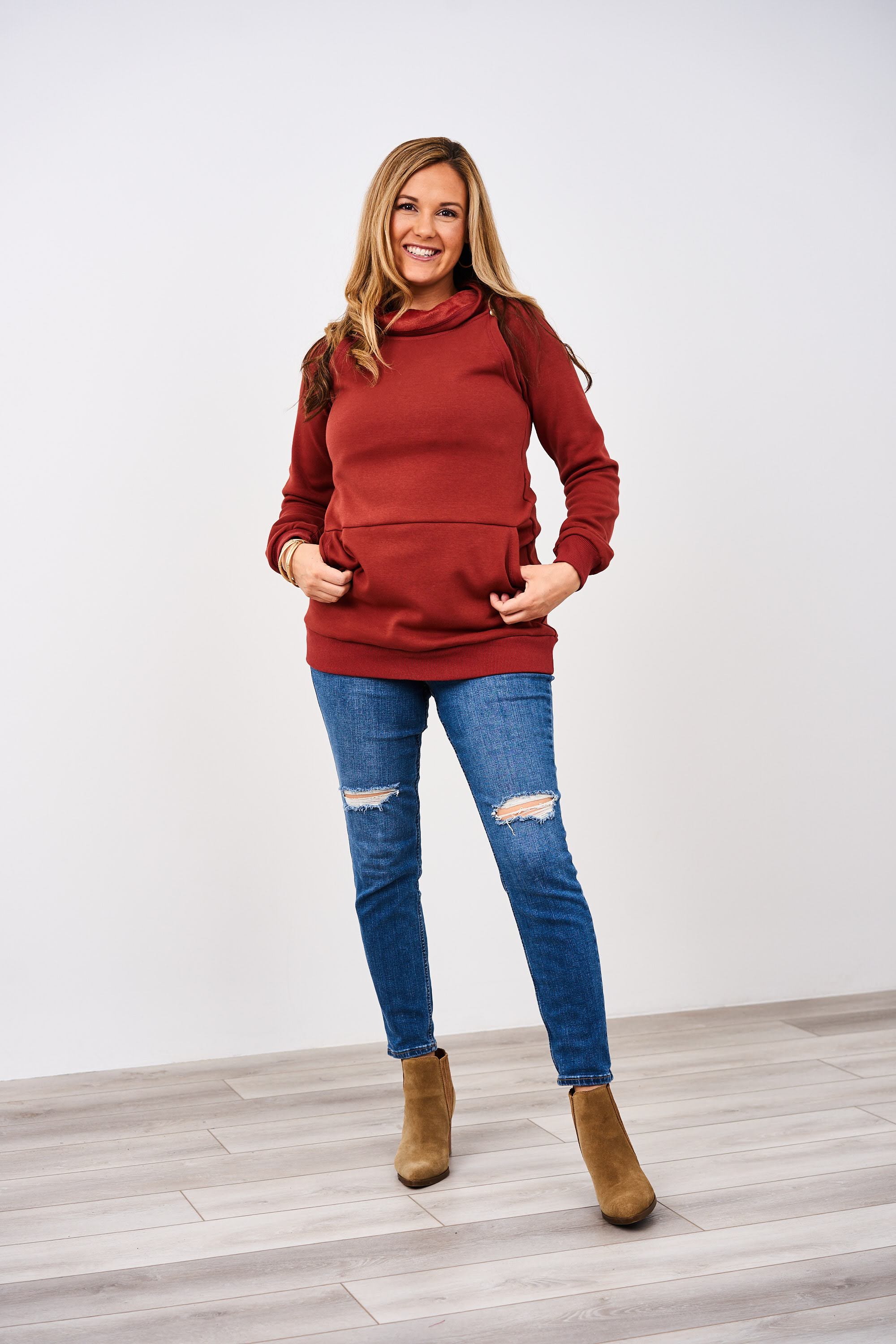 Woman wearing a rust Latched Mama Harbor Snap Nursing Pullover sweater, blue jeans, and brown boots on a white background