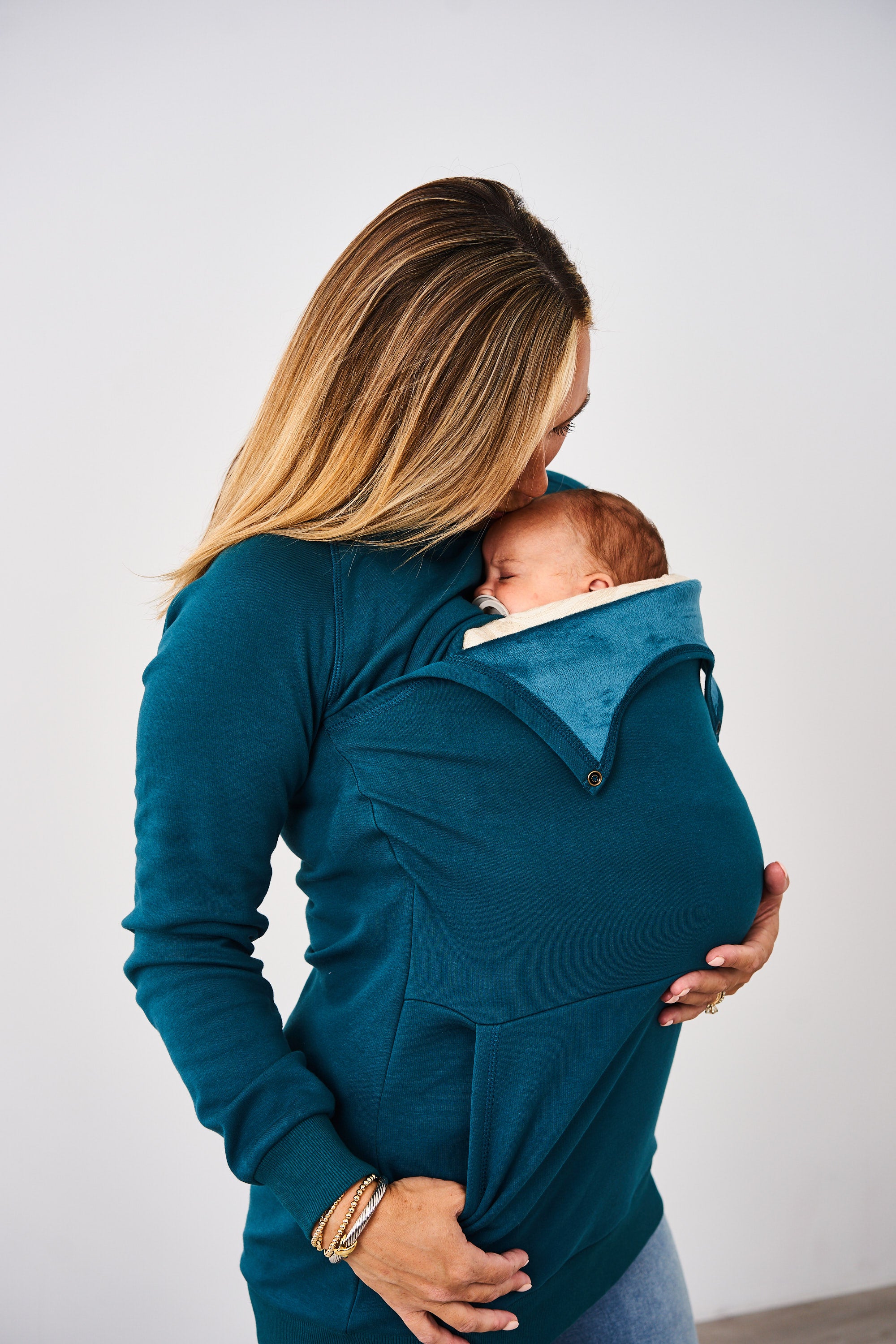 Woman babywearing an infant in a carrier while wearing the Latched Mama Peacock Harbor Snap Nursing Pullover against a white background