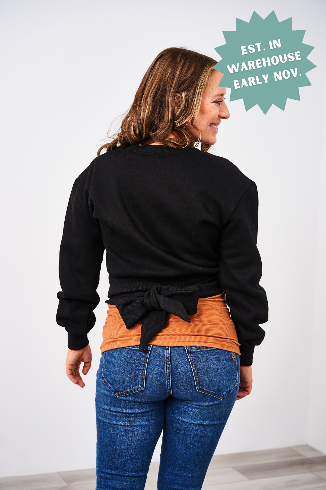 Woman wearing a black sweater tied at the back with an orange shirt underneath, standing against a white wall.