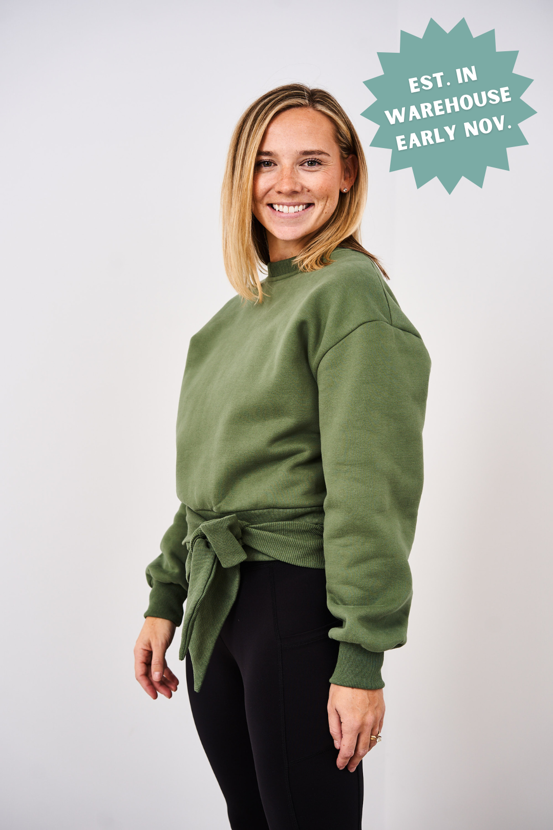 Woman wearing a green sweater with a tie front, standing against a white background.