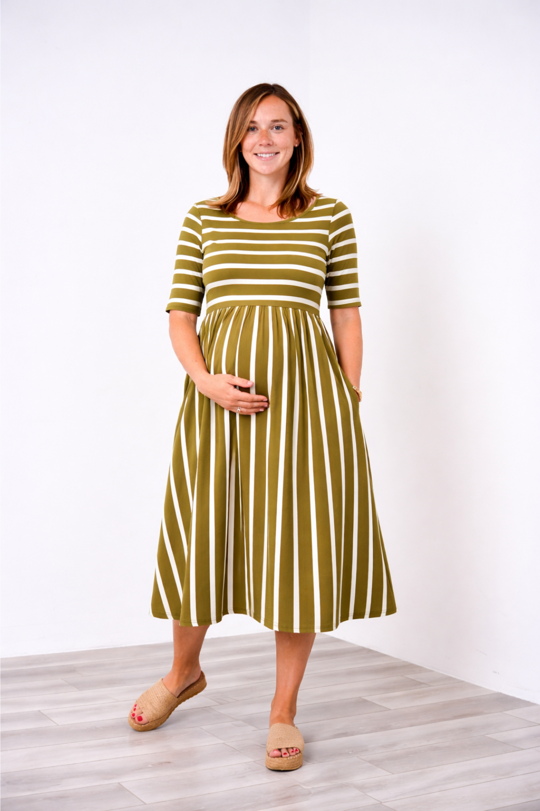 Pregnant woman wearing a green and white striped Latched Mama nursing dress standing against a white background
