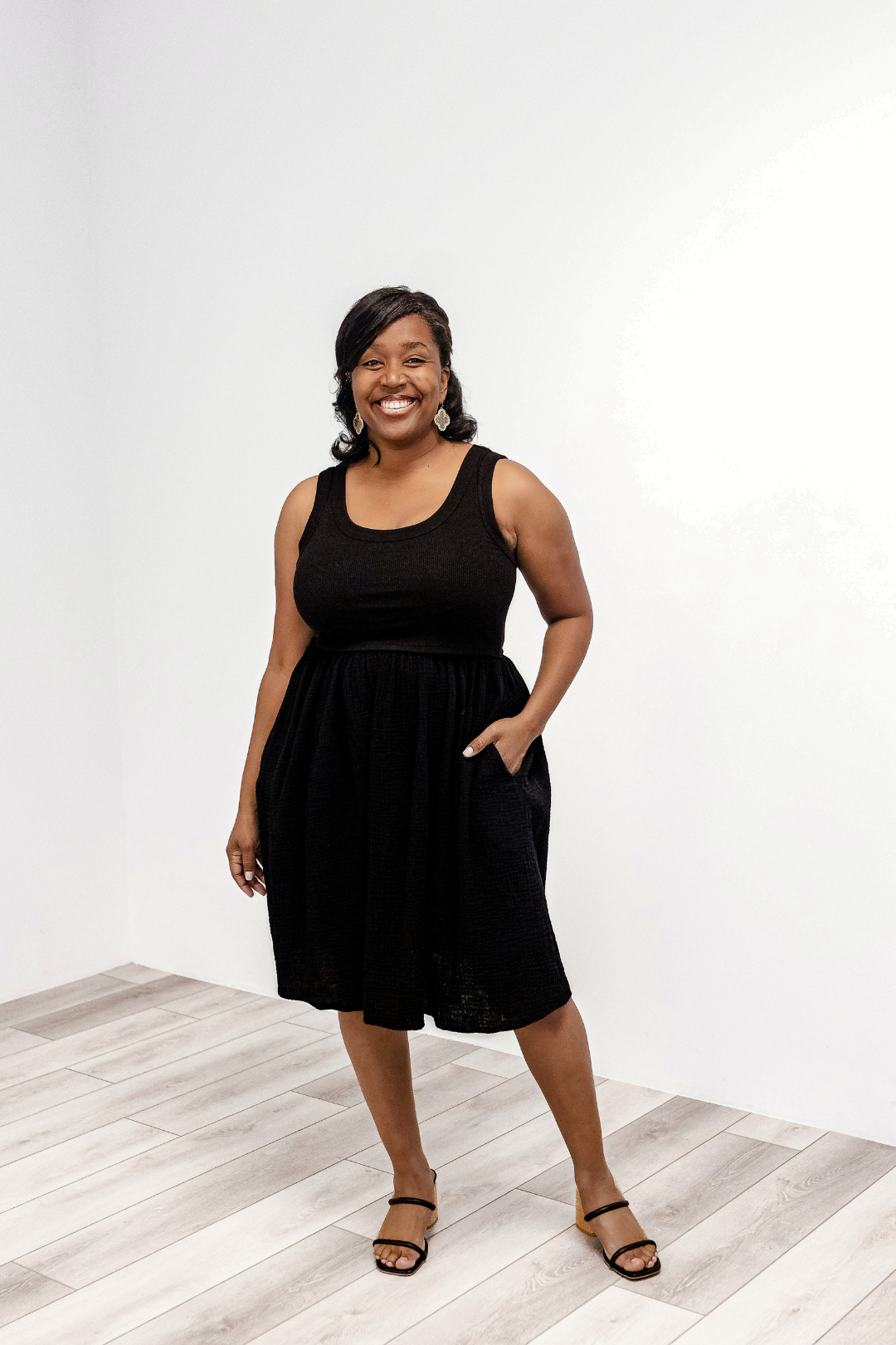 Woman wearing a black sleeveless Latched Mama Breezy Cotton Nursing Dress standing on a light wooden floor with a white background.