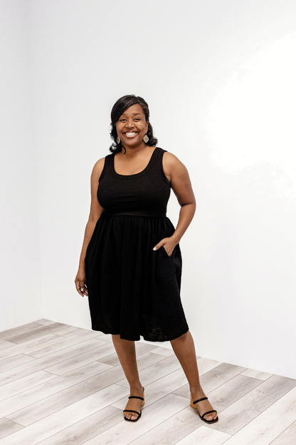 Woman wearing a black sleeveless Latched Mama Breezy Cotton Nursing Dress standing on a light wooden floor with a white background.