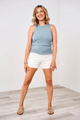 A woman wearing a Latched Mama dusty blue high neck ribbed nursing tank top with white shorts and gold sandals.