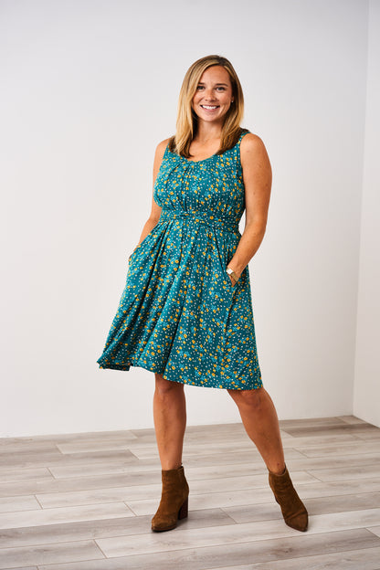 Woman wearing a teal floral dress with brown boots against a white wall.