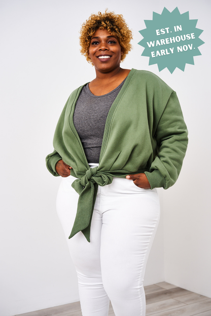 Woman wearing a green cardigan over a gray shirt and white pants, standing against a white background.