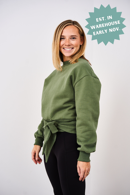 Woman wearing a green sweater with a tie front, standing against a white background.