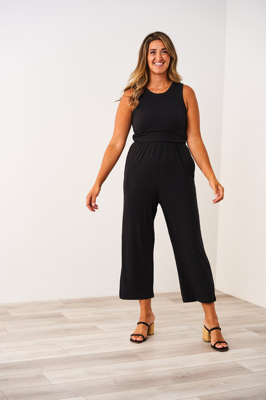 Woman wearing a black Latched Mama nursing jumpsuit standing against a white wall.