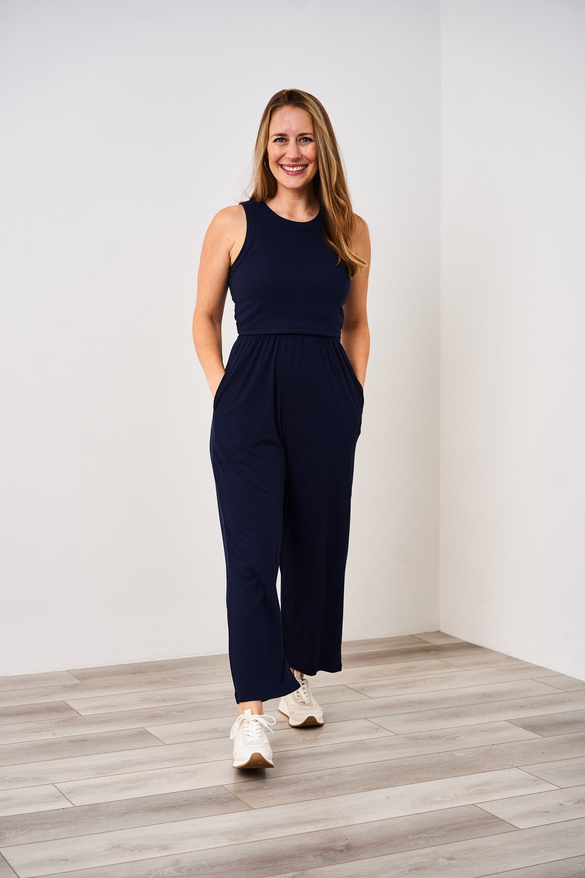 A woman standing in a room wearing a navy blue Latched Mama nursing jumper with white sneakers.